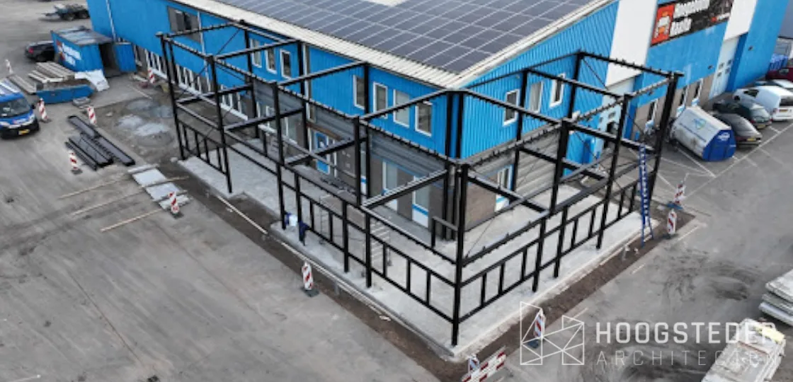 Het hergebruikte stalen frame tijdens de bouw, voor de bestaande blauwe bedrijfshal met zonnepanelen op het dak