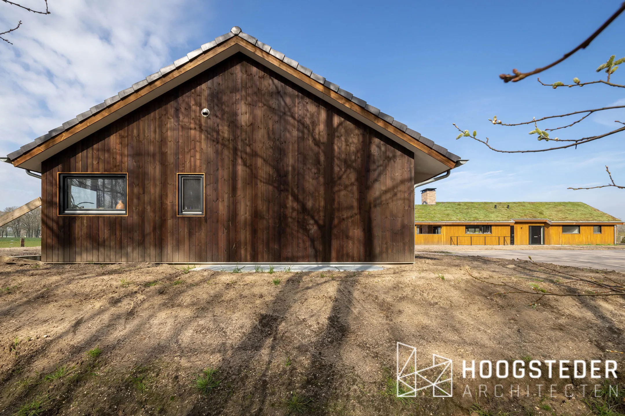 De donkere schuurwoning op de voorgrond en de naturel houten woning met groen dak erachter, samen op één erf in Emst