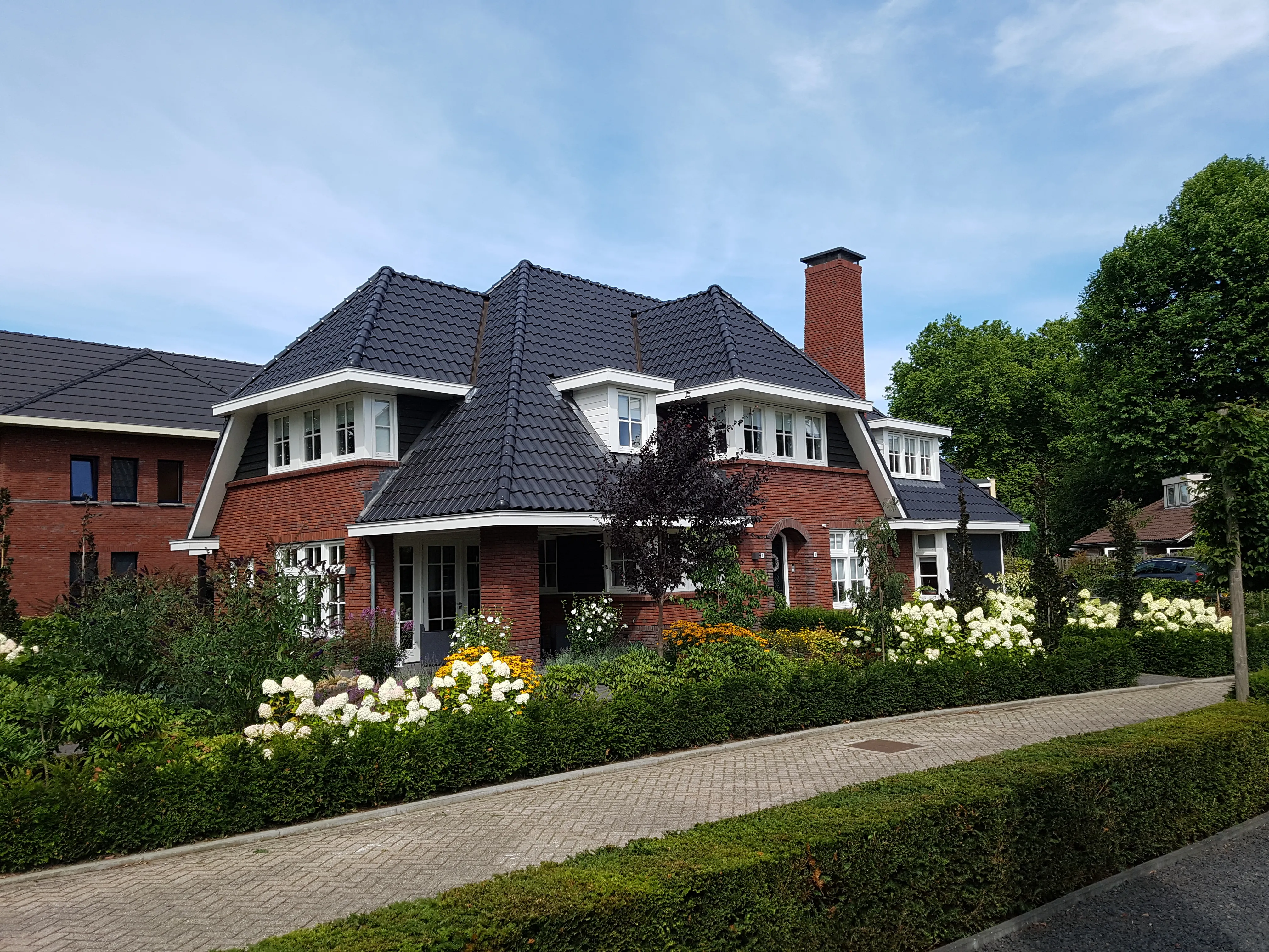 Villa in jaren 30 stijl met rode baksteen, donkere pannen en witte kozijnen, met hortensia's in de voortuin